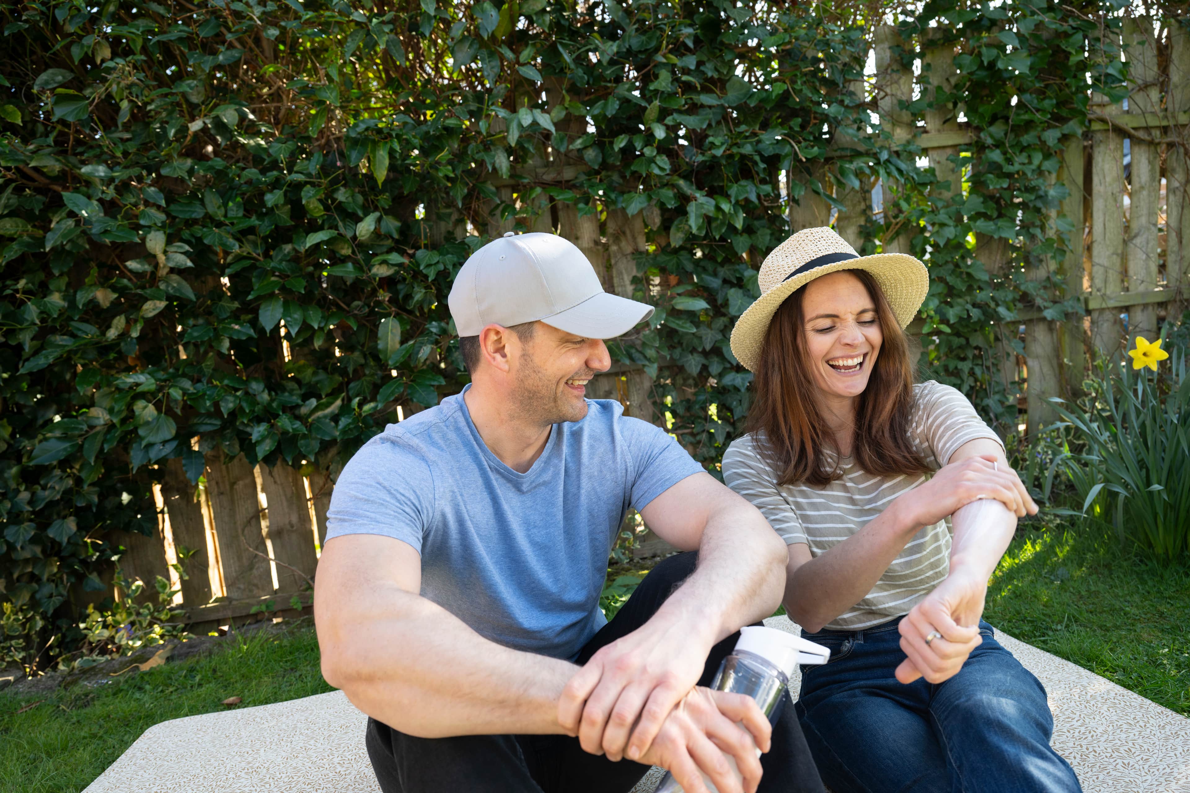 Two adults smiling outdoors in a garden with hats ; one is applying cream to her arm.