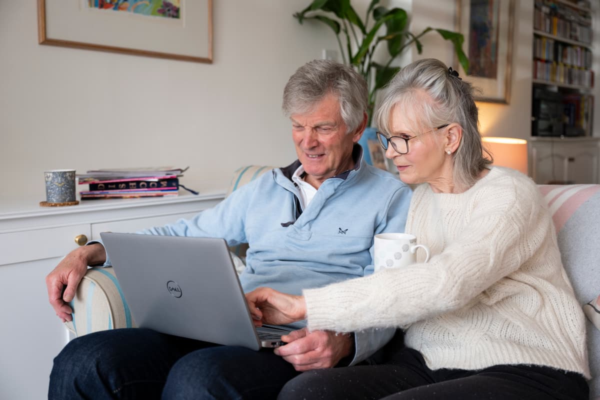 Two older adults look at a laptop screen together in a living room.