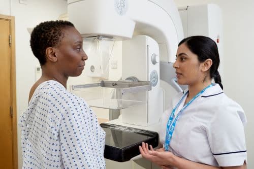 Photo showing a mammographer speaking to a patient, wearing a hospital gown, before a mammogram.